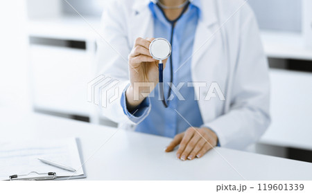 Unknown woman-doctor in blue blouse is holding stethoscope head while sitting at the desk in clinic, close-up. Physician ready to examine and help patient. Medicine concept Unknown woman-doctor in blue blouse is holding stethoscope head while sitting at the desk in clinic, close-up. Physician ready to examine and help patient. Medicine concept 119601339