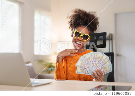 Joyful woman wearing heart-shaped sunglasses, holding a fan of dollar bills, smiling at the camera, expressing financial success and happiness. Joyful woman wearing heart-shaped sunglasses, holding a fan of dollar bills, smiling at the camera, expressing financial success and happiness. 119605083
