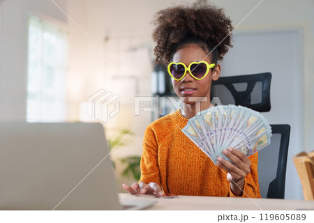 Joyful woman wearing heart-shaped sunglasses, holding a fan of dollar bills, smiling at the camera, expressing financial success and happiness. 119605089