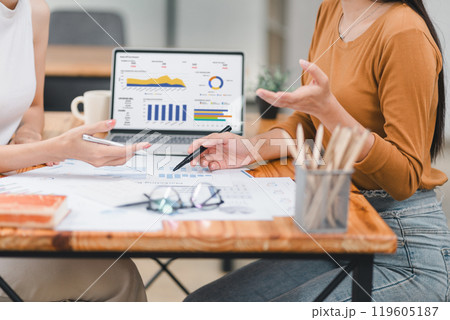 Two professionals analyze business data using laptop displaying sales dashboard and various printed charts on desk. 119605187