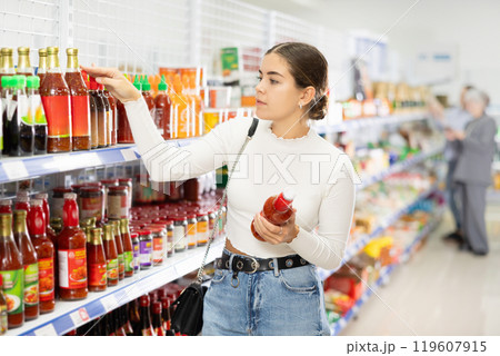 Girl customer is thinking about buying red spicy and unusual sriracha chilli sauce. 119607915