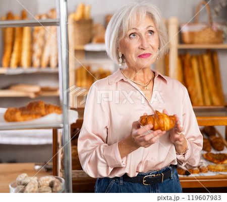 Senior female customer stands with croissant in hands near window of bakery. Senior female customer stands with croissant in hands near window of bakery. 119607983