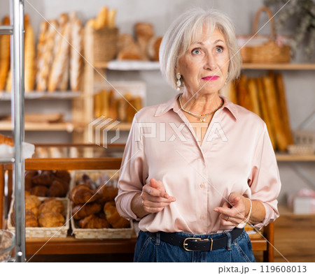 Mature European woman near window of bakery food court store examines assortment 119608013