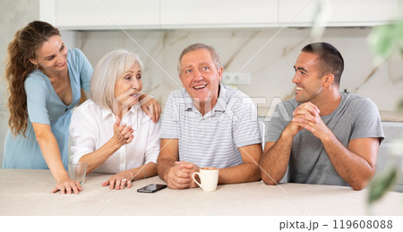Smiling parents and adult children sit at the table with tea and talk about home life 119608088