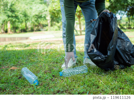 Woman of dedicated volunteers are participating in an environmental cleaning activity in a park. Woman wearing gloves and hold large black bags. Woman collecting trash and plastic bottles in a park. Woman of dedicated volunteers are participating in an environmental cleaning activity in a park. Woman wearing gloves and hold large black bags. Woman collecting trash and plastic bottles in a park. 119611126