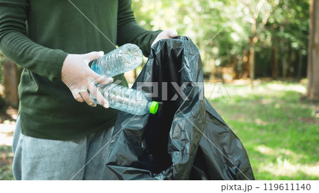 Woman of dedicated volunteers are participating in an environmental cleaning activity in a park. Woman wearing gloves and hold large black bags. Woman collecting trash and plastic bottles in a park. Woman of dedicated volunteers are participating in an environmental cleaning activity in a park. Woman wearing gloves and hold large black bags. Woman collecting trash and plastic bottles in a park. 119611140