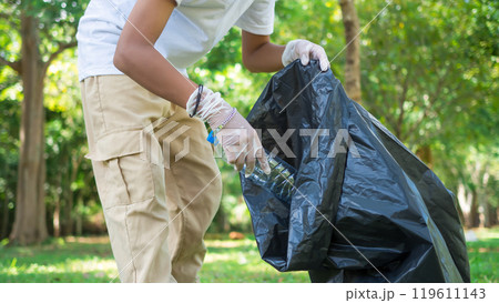 A boy of dedicated volunteers are participating in an environmental cleaning activity in a park. Boy wearing gloves and hold large black bags. Boy collecting trash and plastic bottles in a park. 119611143