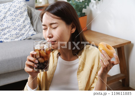 young asian woman enjoy eating sweet donut and drink cola soft drink in living room at home in summer season. young asian woman enjoy eating sweet donut and drink cola soft drink in living room at home in summer season. 119611885