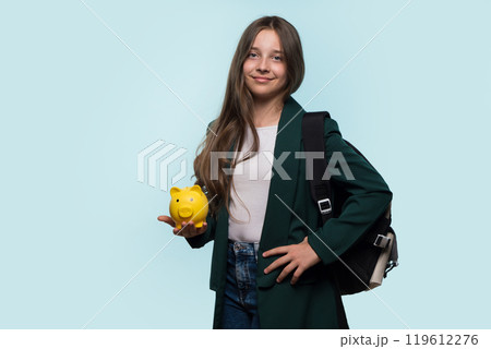Teen girl holding yellow piggy bank, wearing backpack, smiling confidently. 119612276