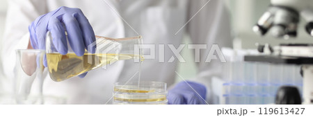 Assistant pours yellow liquid from flask at desk in lab Assistant pours yellow liquid from flask at desk in lab 119613427