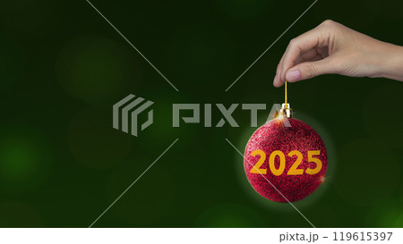 Red Christmas ball with yellow 2025 in woman's hand on green background, horizontal New year style photo banner copy space 119615397