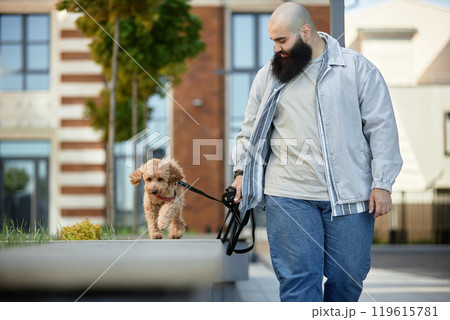 Man walking his dog while holding leash, enjoying outdoor stroll beside modern city buildings without looking at camera 119615781