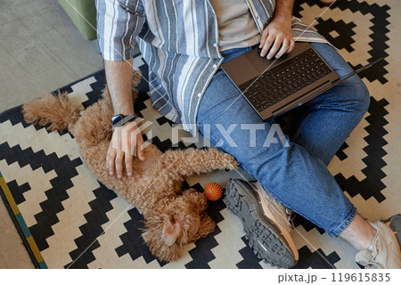 Casual setting showing person using laptop while sitting on floor with fluffy dog lying beside. Person wearing striped shirt and blue jeans, and seated on patterned rug 119615835