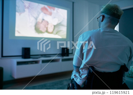 Person sitting on chair in living room watching movie on large projector screen with surround sound system and white shelving unit in background Person sitting on chair in living room watching movie on large projector screen with surround sound system and white shelving unit in background 119615921