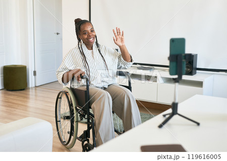 Smiling African American woman in wheelchair holding smartphone on stand in modern living room setting with white furniture and hardwood flooring Smiling African American woman in wheelchair holding smartphone on stand in modern living room setting with white furniture and hardwood flooring 119616005