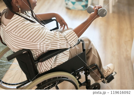Person in wheelchair exercising with dumbbell in cozy interior. Hand has relaxed grip on weight while focused on activity, creating positive, motivated atmosphere 119616092