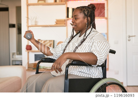 Black person sitting in wheelchair and exercising with dumbbell in hand while surrounded by home environment, showcasing active and positive lifestyle 119616093