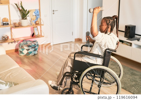 Person in wheelchair lifting dumbbells in home setting filled with various household items. Decor including plants and decor pieces surrounds the area without using professional equipment 119616094
