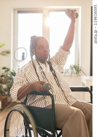 Young African American woman wheelchair user engaging in exercise routine with dumbbell in a home setting filled with natural light 119616095