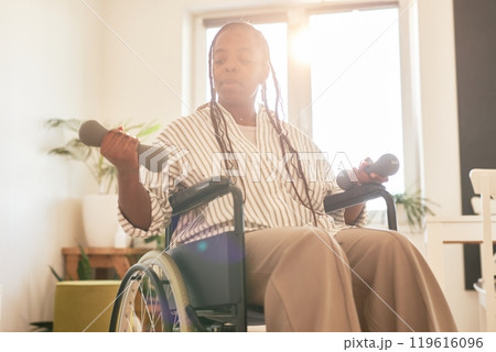 African American woman in wheelchair lifting dumbbells with focused expression in brightly lit room during day. Plant and shelf visible in background creating a homely atmosphere 119616096