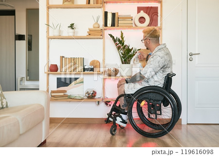 Man with tattoos in wheelchair sitting near bookshelf with decorative items in modern living room with wooden floor and white walls, looking sideways Man with tattoos in wheelchair sitting near bookshelf with decorative items in modern living room with wooden floor and white walls, looking sideways 119616098