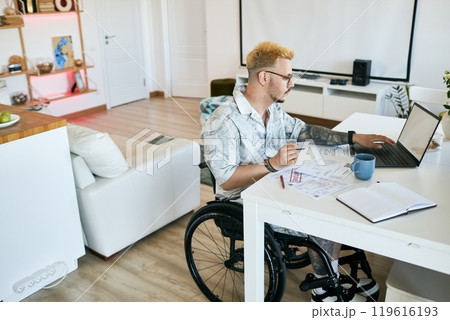 Man sitting in his wheelchair, working on laptop at home office desk, surrounded by papers, notes, and coffee cup. Modern, clean living space visible with shelves and decor Man sitting in his wheelchair, working on laptop at home office desk, surrounded by papers, notes, and coffee cup. Modern, clean living space visible with shelves and decor 119616193
