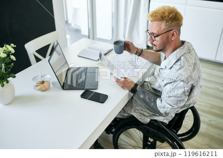 Man in wheelchair reading documents and holding mug while working at home office table showing focus and determination using technology and devices 119616211