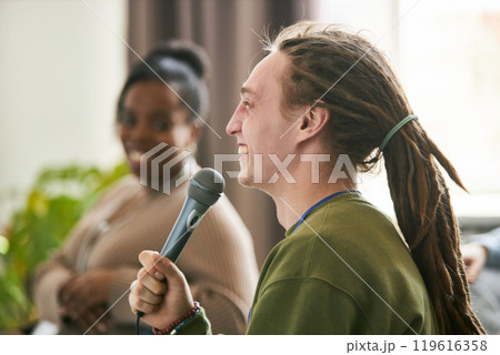 Man with dreadlocks happily speaking into a microphone, with another person blurred in the background creating a lively and interactive atmosphere Man with dreadlocks happily speaking into a microphone, with another person blurred in the background creating a lively and interactive atmosphere 119616358