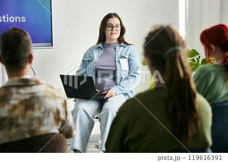 Woman with laptop presenting to audience in casual business setting. Group paying attention while listening to presentation, plant and screen seen in background 119616391