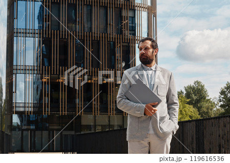 Businessman holding tablet while standing in front of modern office building on a clear day wearing light-colored suit and looking thoughtfully into distance 119616536