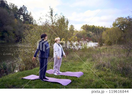 middle-aged woman teaches her old mother somatic exercises and yoga to maintain health by the river in autumn 119616844