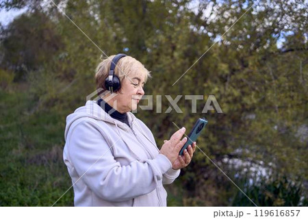 elderly woman switches the music in her wireless headphones using her phone during a morning run in the autumn park 119616857