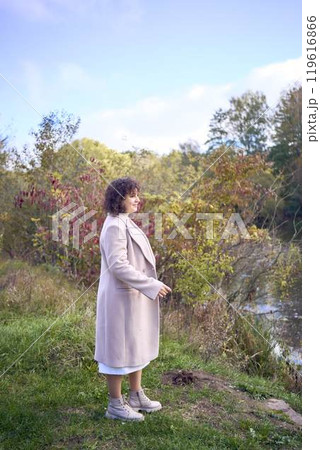 middle-aged woman with dark curly hair in light beige suit and coat enjoying nature in autumn forest 119616866