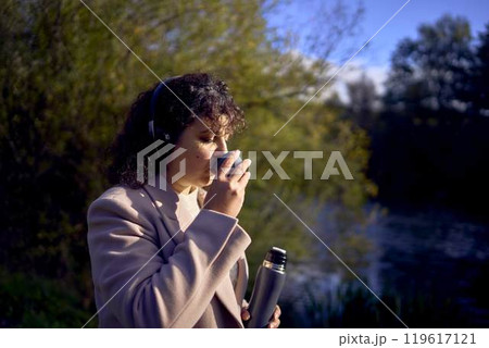 middle-aged woman in wireless headphones drinking coffee, tea from a thermos on a walk in the autumn forest middle-aged woman in wireless headphones drinking coffee, tea from a thermos on a walk in the autumn forest 119617121