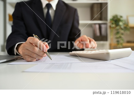 Focused accountant in black suit analyzing financial data at office desk Focused accountant in black suit analyzing financial data at office desk 119618010
