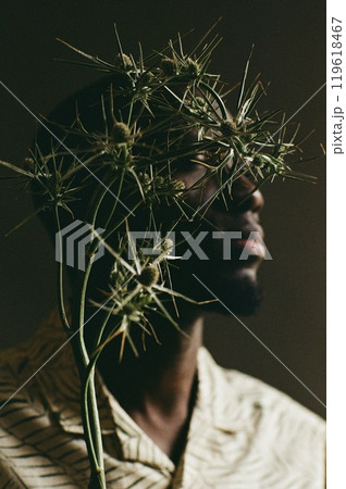 Artistic close up portrait of young African American man with eryngium plant hiding face shot on film with grain Artistic close up portrait of young African American man with eryngium plant hiding face shot on film with grain 119618467