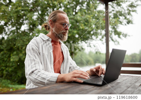 Side view portrait of bearded adult man using computer while working outdoors at terrace by beautiful lake in nature copy space 119618600