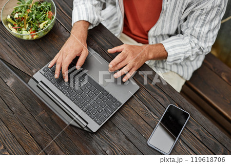 Top view closeup of unrecognizable adult man typing on laptop keyboard while working at table in outdoor cafe or terrace 119618706
