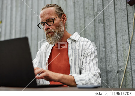 Minimal portrait of bearded adult man with suntan and glasses using laptop outdoors while sitting at table with metal wall in background copy space 119618707