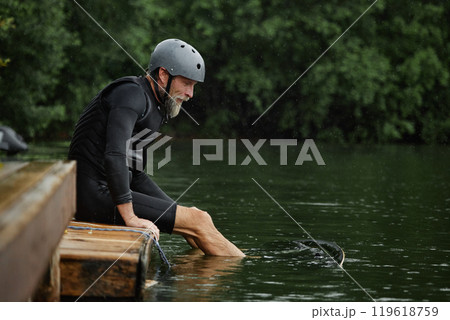 Side view portrait of active senior man sitting on pier by ake with feet in water and wearing protective gear for sports copy space 119618759