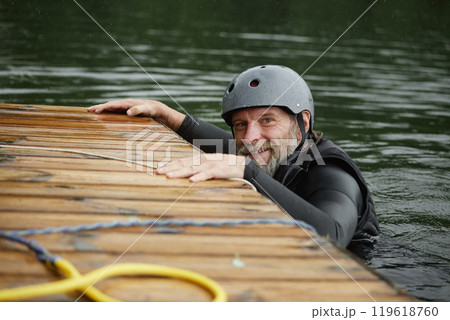 Portrait of smiling bearded man in lake water holding onto wooden pier and smiling at camera practicing water sports copy space 119618760