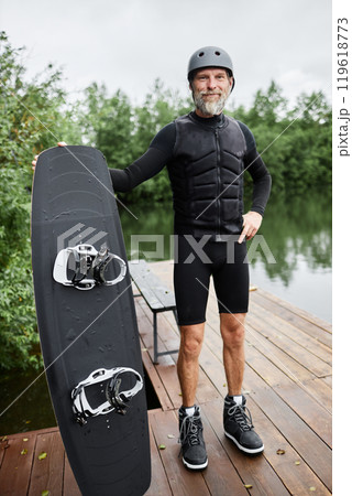 Vertical full length portrait of bearded senior man wearing wakeboarding gear and holding board smiling at camera standing on pier by lake outdoors 119618773