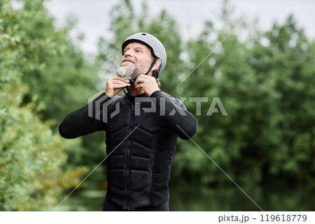 Waist up portrait of bearded senior man putting on wetsuit and helmet ready to enjoy water sports by lake in forest copy space Waist up portrait of bearded senior man putting on wetsuit and helmet ready to enjoy water sports by lake in forest copy space 119618779