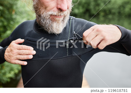 Close up of bearded mature man putting on wetsuit by lake and closing velcro fastenings getting ready for diving Close up of bearded mature man putting on wetsuit by lake and closing velcro fastenings getting ready for diving 119618781