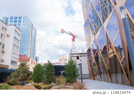 contemporary urban area features newly built glass fronted structure, surrounded by greenery, with construction equipment crane working in background under overcast sky 119619474