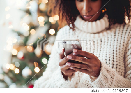 Black woman in cozy sweater texting on smartphone against festive background. Christmas, New Year concept 119619807