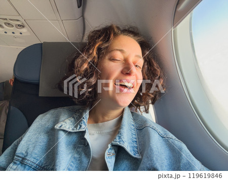 Happy passenger young woman is flying in plane. Smiling girl sitting in airplane near window going on trip vacation travel. Traveling female inside plane enjoying flight. Traveling girl Happy passenger young woman is flying in plane. Smiling girl sitting in airplane near window going on trip vacation travel. Traveling female inside plane enjoying flight. Traveling girl 119619846