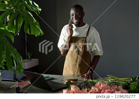 Waist up portrait of smiling African American man speaking on phone and looking at laptop screen while managing flower shop with elegant dark setting Waist up portrait of smiling African American man speaking on phone and looking at laptop screen while managing flower shop with elegant dark setting 119620277