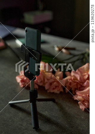 Vertical close up of smartphone on tripod stand and flowers laid out on table with low lighting Vertical close up of smartphone on tripod stand and flowers laid out on table with low lighting 119620278