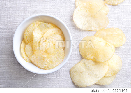 Salted potato chips in a white bowl on linen fabric. Fresh and ready to eat crisps, slim slices of deep fried potato, made from tubers of the root vegetable Solanum tuberosum. Close-up, from above. Salted potato chips in a white bowl on linen fabric. Fresh and ready to eat crisps, slim slices of deep fried potato, made from tubers of the root vegetable Solanum tuberosum. Close-up, from above. 119621161